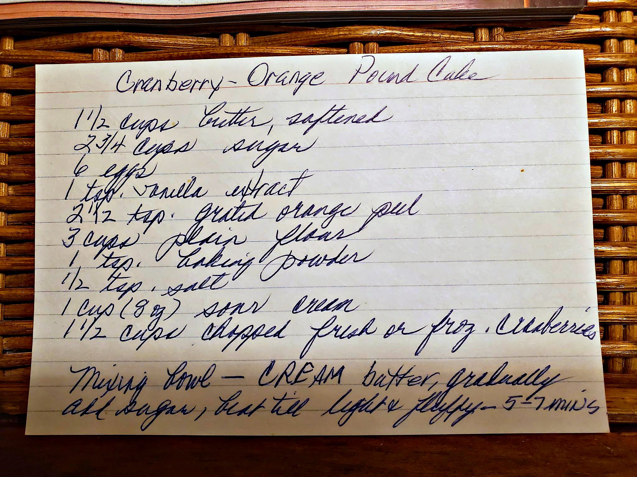 Hand written recipe card for cranberry orange pound cake recipe card sitting on wicker basket.