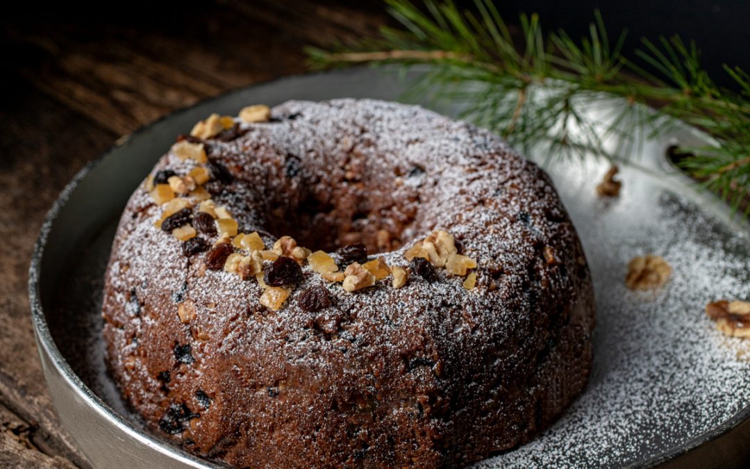Fruit Cake Bundt cake on a silver platter with a pine bow in the background. Decorated with raisins, candied orange peel, and walnuts.